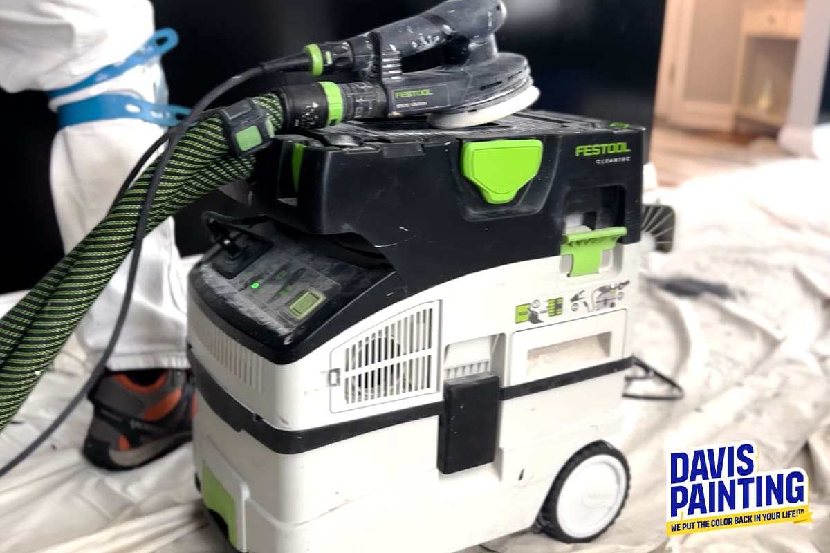 A Festool dust extractor is connected to a sander. A worker in white overalls and a blue safety harness is partially visible behind the equipment. The machine is on a white drop cloth in a room with light-colored furniture. The Davis Painting logo is in the bottom right corner.