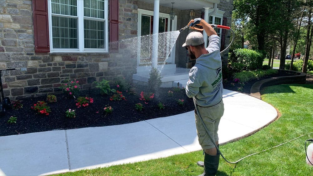 A person wearing a cap and boots is holding a hose, watering plants in a well-maintained garden with colorful flowers. The garden is in front of a house with a stone exterior, large windows, and a white porch. Nearby, the smell of fresh interior painting hints at recent home updates. A sidewalk curves in front of the garden.
