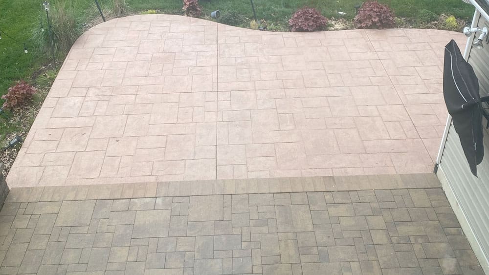 A backyard patio featuring interlocking stone pavers in two distinct colors and patterns: beige with a grid design and a larger pinkish section with a more abstract design. Landscaping elements like grass, shrubs, and flowers border the patio, creating an inviting space that complements the home's interior painting.