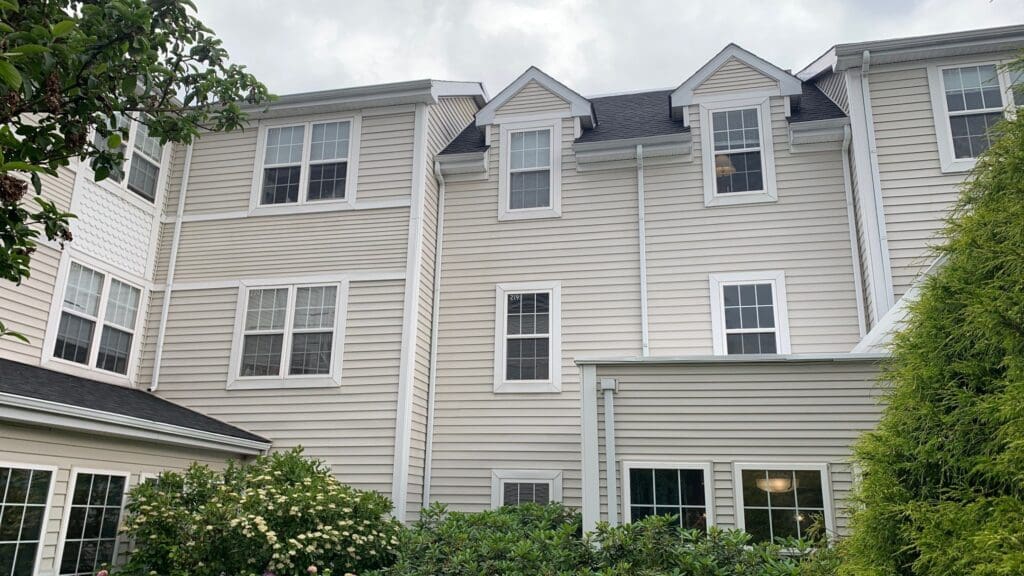 A multi-story beige house with white trim is shown. It has several windows of varying sizes and styles, including double-hung and dormer windows. Green bushes and trees surround the base of the house. The interior painting complements its exterior charm perfectly, even on an overcast day with gray clouds.