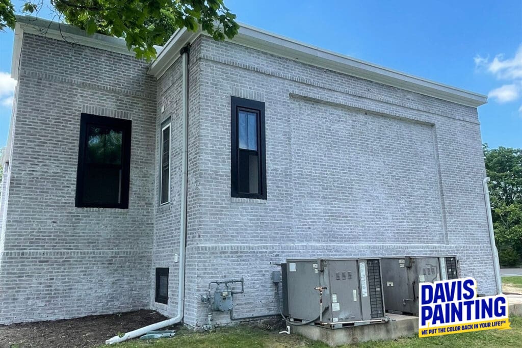 A newly painted white brick building with black window frames and HVAC units on the green grass alongside it. The building has a clean, fresh look under a clear blue sky, reminiscent of the meticulous care seen in a church painting. A "Davis Painting" logo stating "We Put the Color Back in Your Life!" is in the bottom right corner.