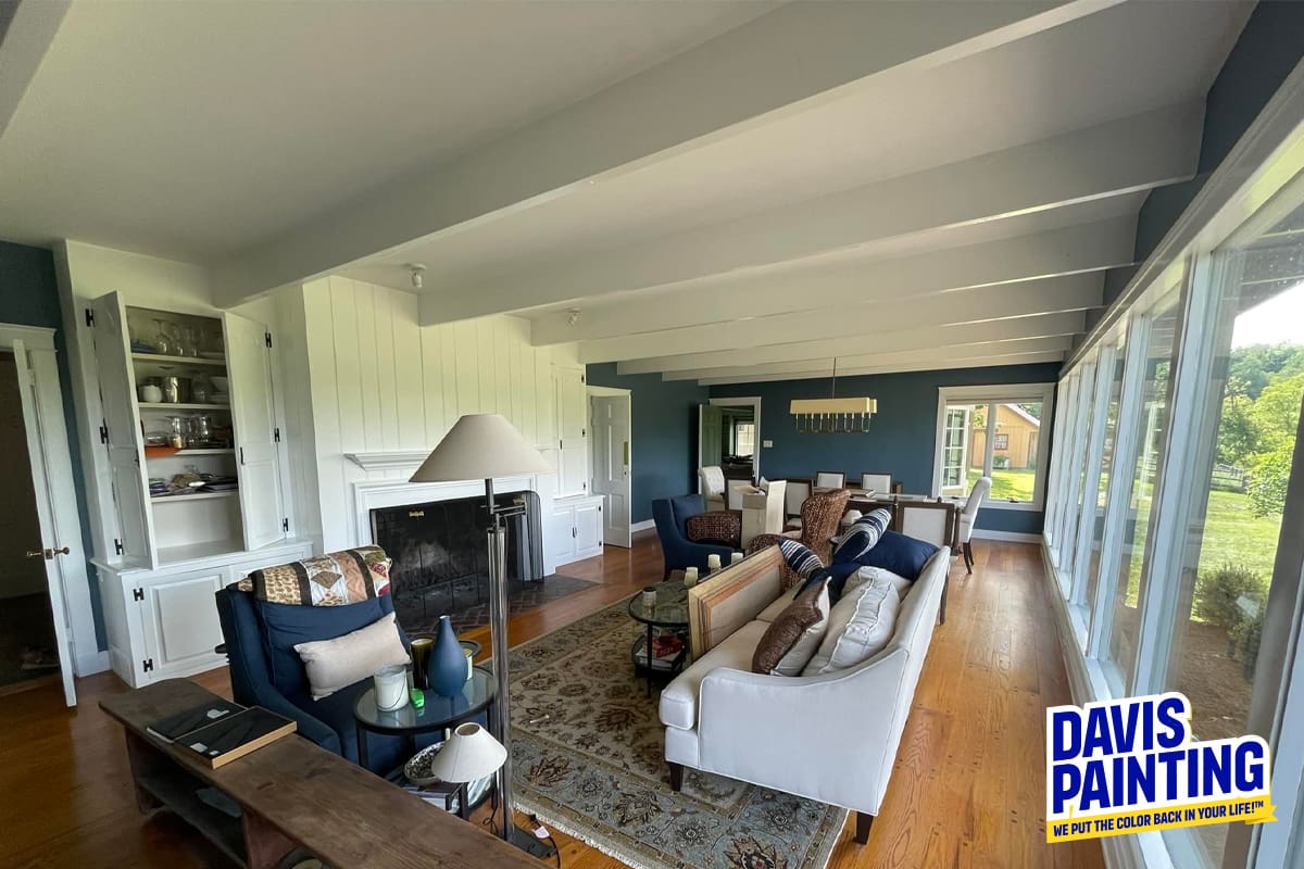 A spacious, modern living room with large windows offering a scenic view showcases exquisite interior design. The room features white walls, dark blue accents, a cozy fireplace, comfortable sofas, and a wooden dining set. The logo "Davis Painting" is displayed in the bottom right corner.