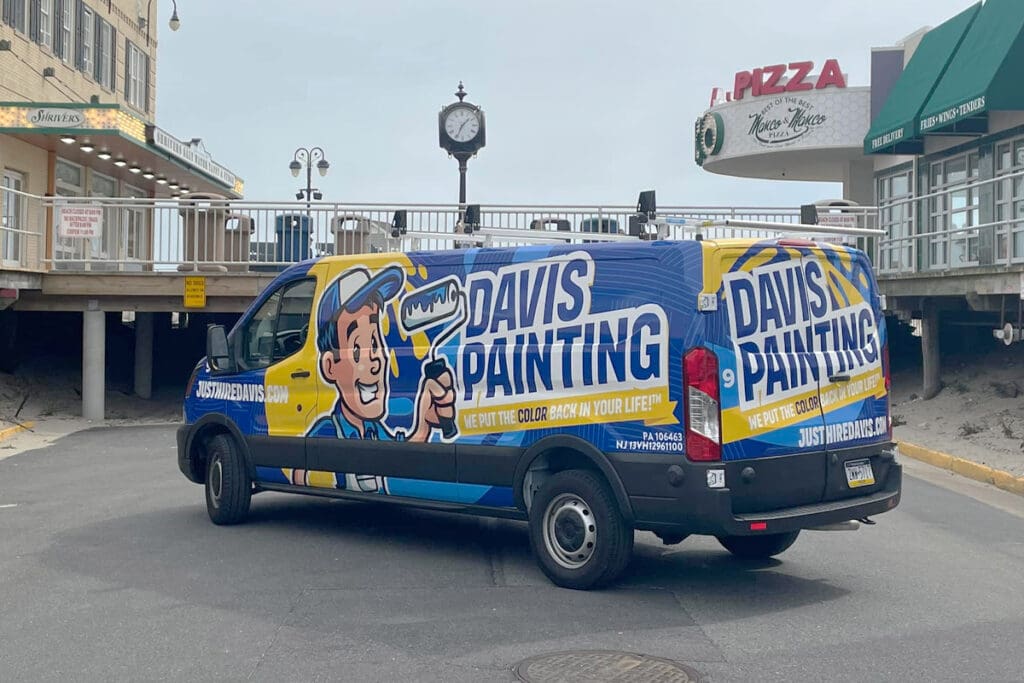 A van with colorful graphics displaying "Davis Painting" and a cartoon painter is parked on a street near a boardwalk dotted with shops and a clock. The website justwetyourbrush.com is also visible, inviting inquiries for painting careers.