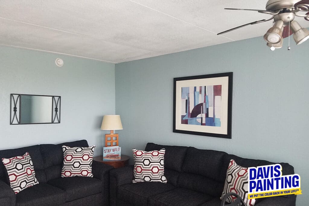 A living room with light blue walls features two dark sofas adorned with patterned cushions. A modern abstract painting, reminiscent of a church painting, and a rectangular mirror are mounted on the walls. A lamp and decor books sit on a side table beneath the ceiling fan. "Davis Painting" logo in corner.