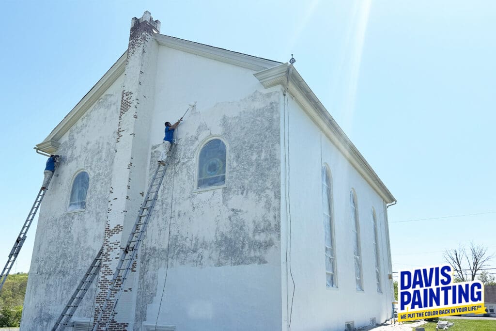 Two painters on ladders work on the exterior wall of a large, old church, undertaking a careful church painting and restoration. The wall is partly weathered and partly freshly painted white. A "Davis Painting" logo appears at the bottom right.