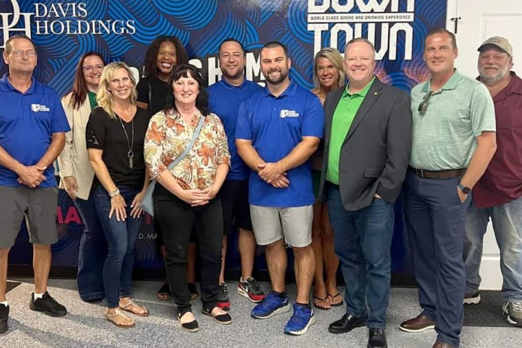 A group of eleven people, both men and women, stand indoors smiling at the camera. Some wear business attire while others are in casual clothes. A blue and white Davis Holdings banner promoting cabinet painting is visible in the background.