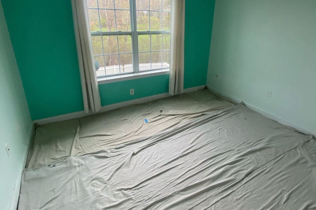 An empty room with teal and light green walls, a large window with white curtains, and floors covered with drop cloths, reflecting current interior painting trends in preparation for a fresh renovation.