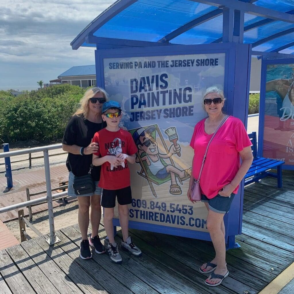 Three people stand and smile next to a bus stop poster advertising Davis Painting Jersey Shore. The group is on the Wildwoods boardwalk by the beach, with greenery and a blue sky in the background.