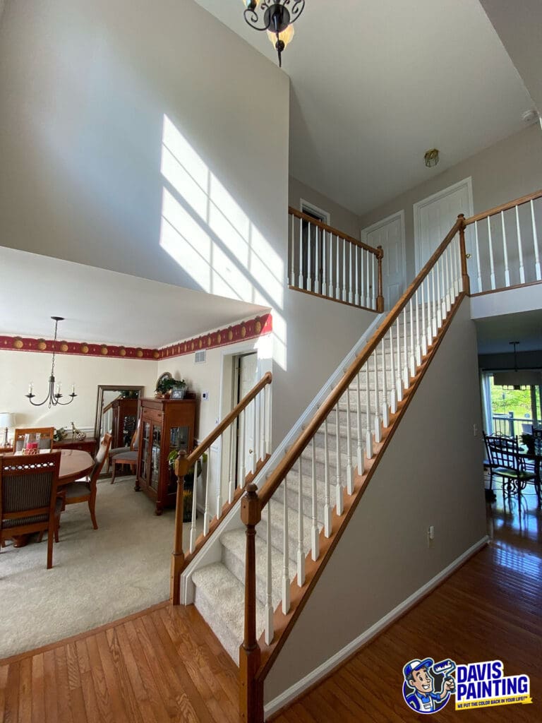 A well-lit home interior with a carpeted staircase, wooden railing, and beige walls enhanced by professional interior painting. The dining room is visible on the left, and the Davis Painting logo appears in the lower right corner.