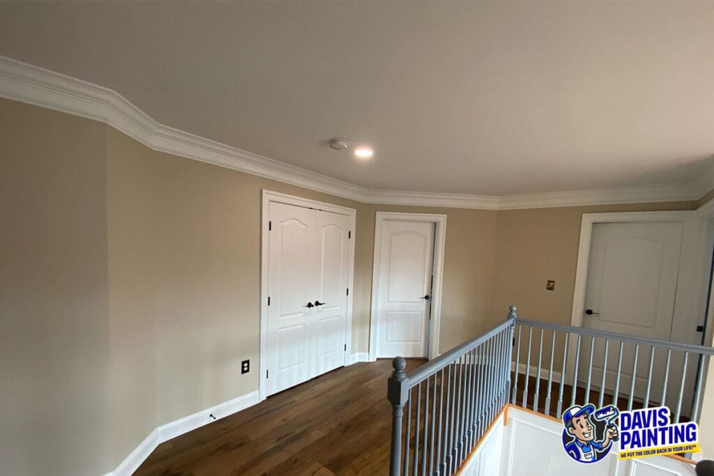 A freshly painted room with beige walls, white trim, and crown molding showcases expert interior painting. The space features two white double doors, wood flooring, and a staircase with gray railings. The “Davis Painting” logo appears in the bottom right corner.