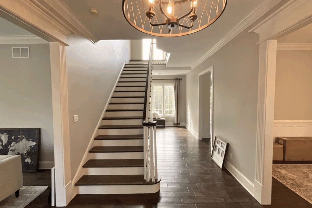 18 A bright, modern entryway with dark wood floors, a staircase with white railings, a chandelier overhead, and large windows at the back letting in natural light. Framed pictures and boxes rest against the walls.