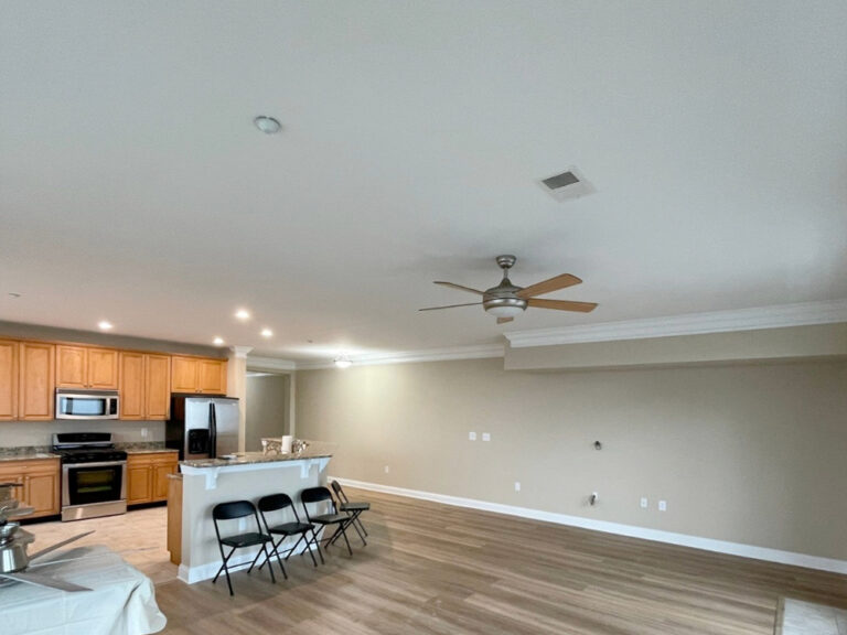 A spacious, unfurnished living area with wood floors, a ceiling fan, and beige walls. An open kitchen with wooden cabinets and stainless steel appliances is visible, along with three black folding chairs by the counter.