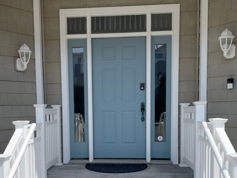 A light blue front door with glass sidelights and a transom window, flanked by white railings and two white wall lanterns, leading onto a porch with gray siding.
