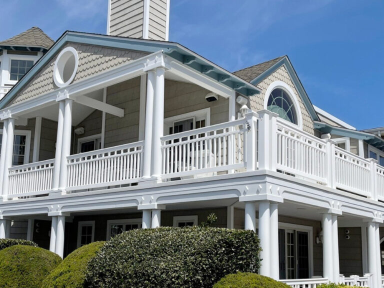 A two-story house with white railings, columns, and tan siding, featuring a large wraparound porch and round windows; trimmed bushes are in the foreground under a clear blue sky.