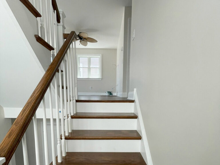 A staircase with wooden steps and white railings leads up to a hallway with a ceiling fan and a window letting in natural light. A small blue paint tray and tools sit on the floor at the top of the stairs.