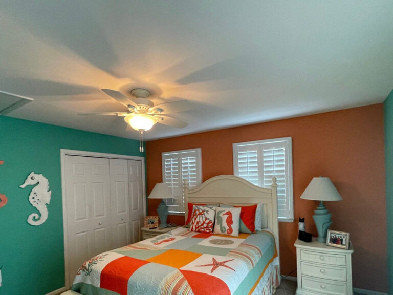 A beach-themed bedroom with teal and orange walls, a bed with colorful sea life bedding, two white nightstands with lamps, shuttered windows, and a ceiling fan with a light. A seahorse decoration is on the wall.