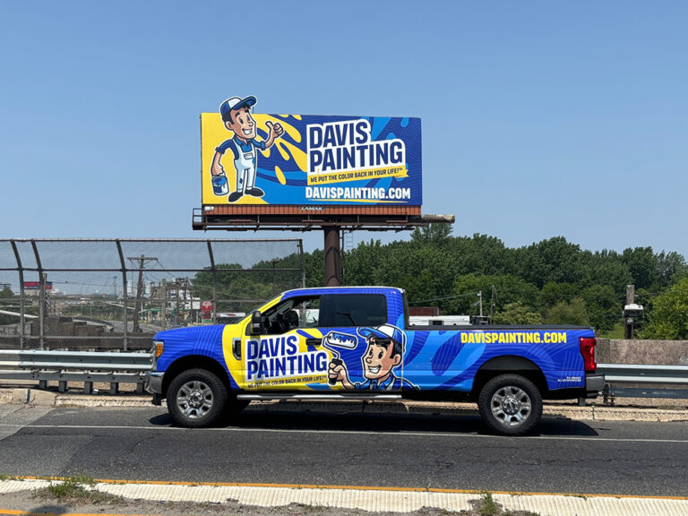 A blue pickup truck with a "Davis Painting" logo is parked in front of a matching "Davis Painting" billboard along a highway. Both feature a cartoon painter holding a brush and website davispainting.com. Trees and road visible.