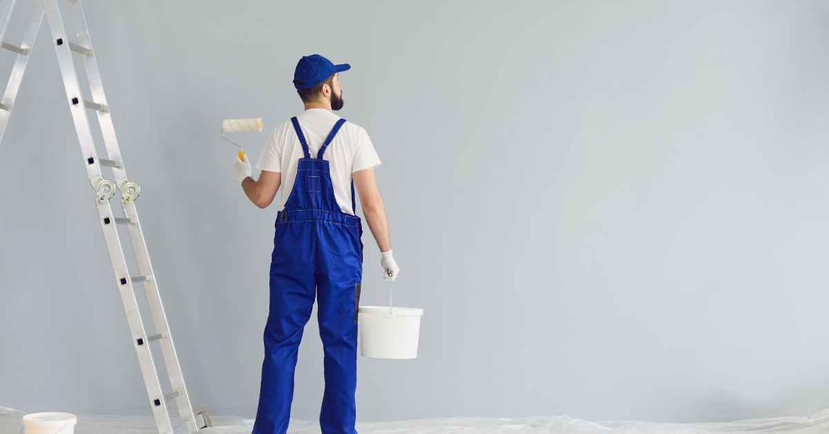 A person in blue overalls and a cap stands by a ladder, holding a paint roller and a white paint bucket, facing a blank light gray wall, preparing to paint. Plastic sheeting covers the floor.