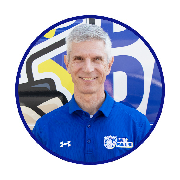 A smiling man named Chuck with short gray hair wears a blue Davis Painting polo shirt and stands in front of a colorful painted backdrop.