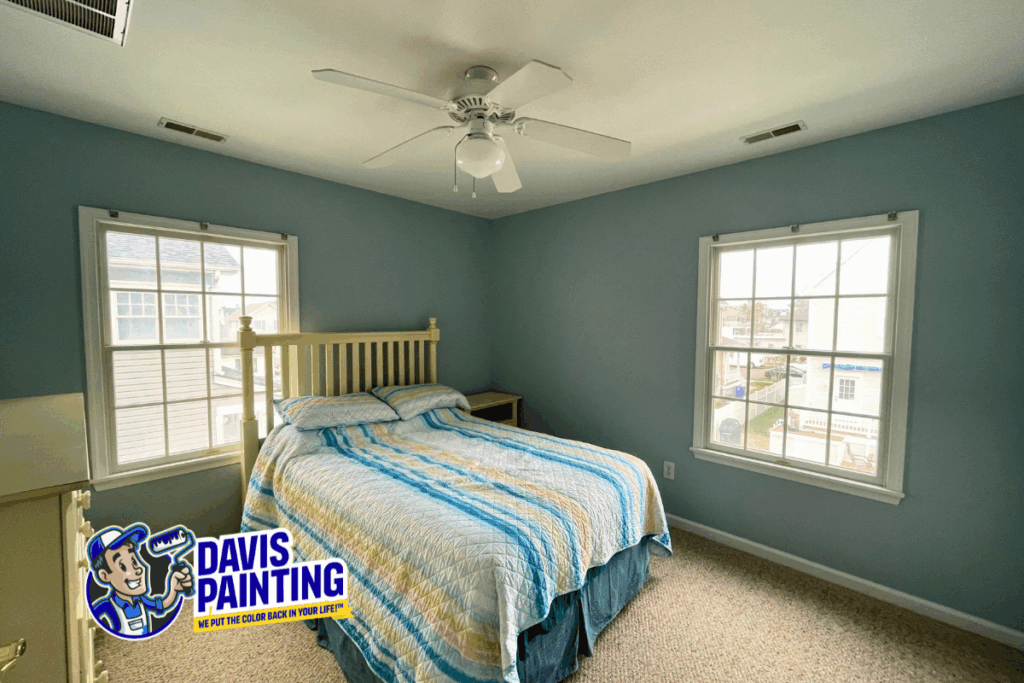 A neatly made bed with striped bedding sits between two large windows in a bright blue painted bedroom. A logo in the corner reads "Davis Painting" with a cartoon painter holding a roller.