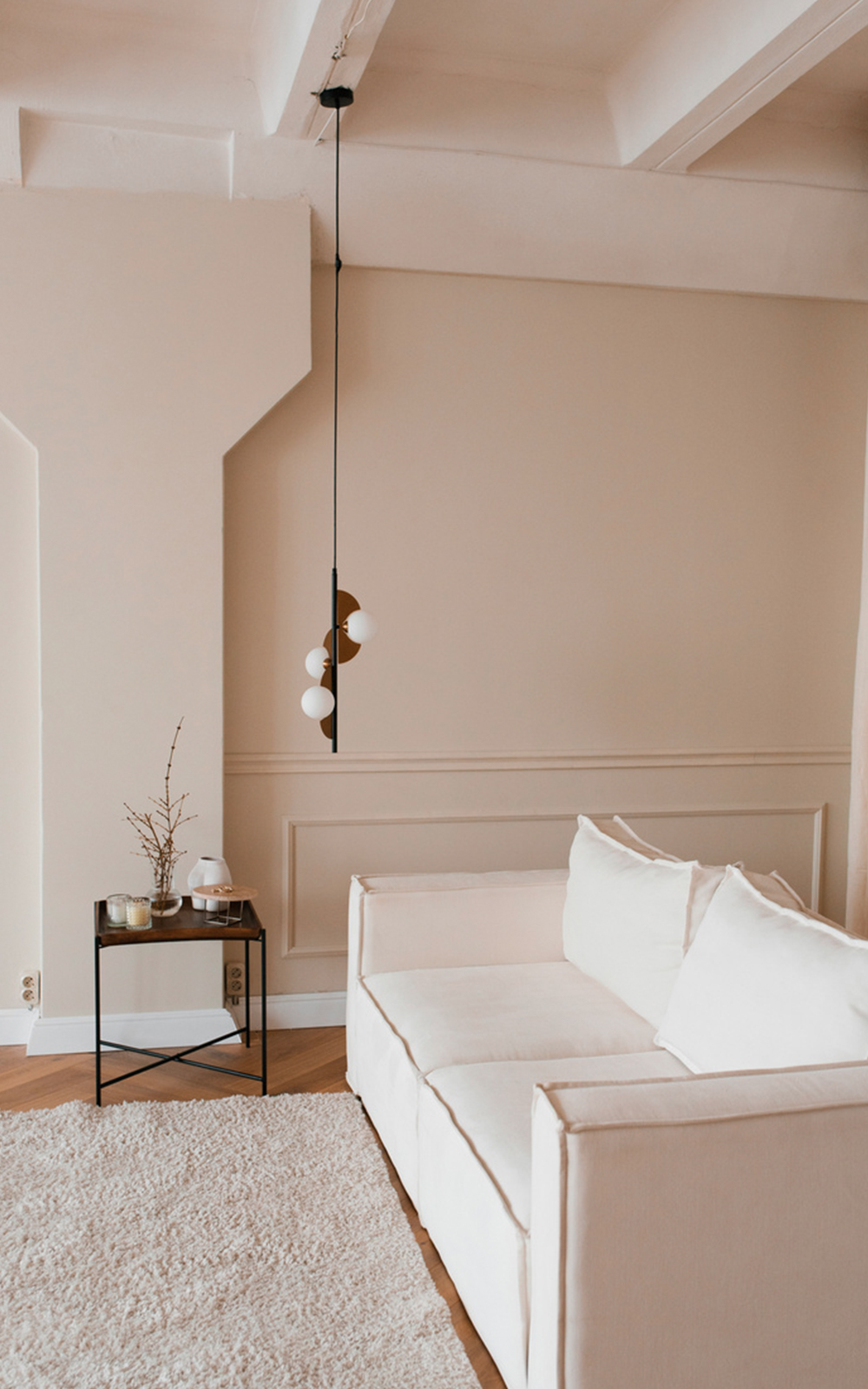 Minimalist living room design featuring a cream-colored sofa, white cushions, a textured rug, a small side table with decor, and a modern pendant light hanging from the ceiling against beige walls.