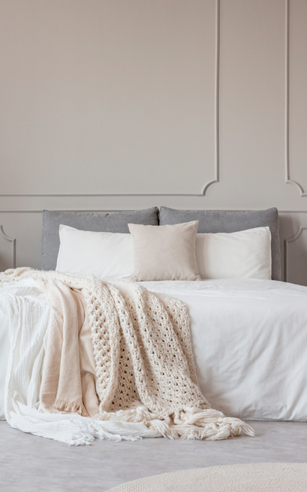 A neatly made bed with white linens, a cream throw blanket, and three pillows—two white and one beige—showcases elegant design against a light gray upholstered headboard and a beige wall with sophisticated molding.