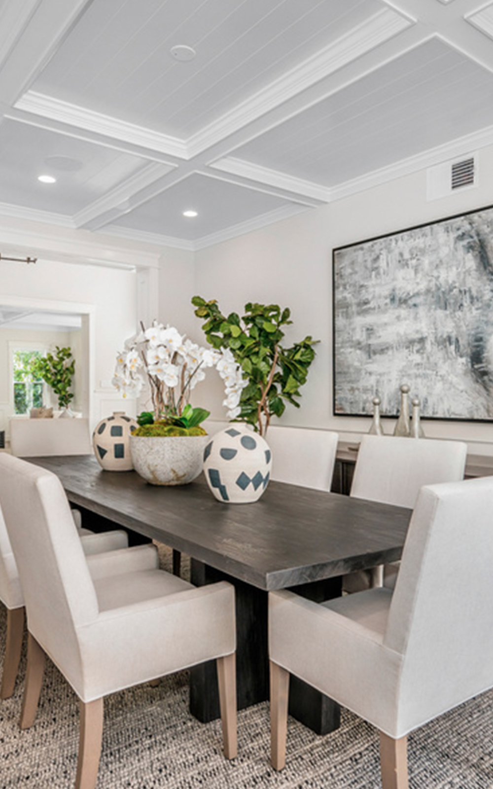 Modern dining room with a black rectangular table, six beige upholstered chairs, decorative vases, white orchids, a green plant centerpiece, abstract wall art, and neutral-toned decor in a bright setting that highlights elegant design.