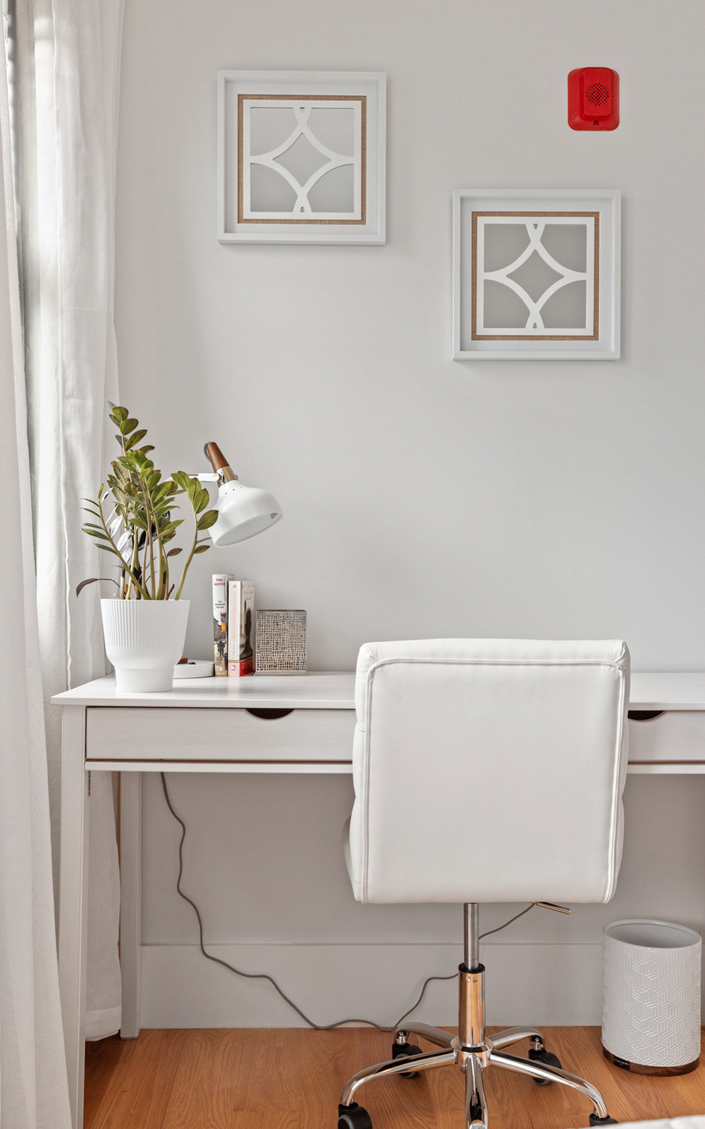 A tidy white desk with a white chair, a potted plant, desk lamp, and books showcases minimalist design against a white wall with two decorative framed mirrors and a red smoke detector above. A trash bin sits neatly under the desk on a wooden floor.