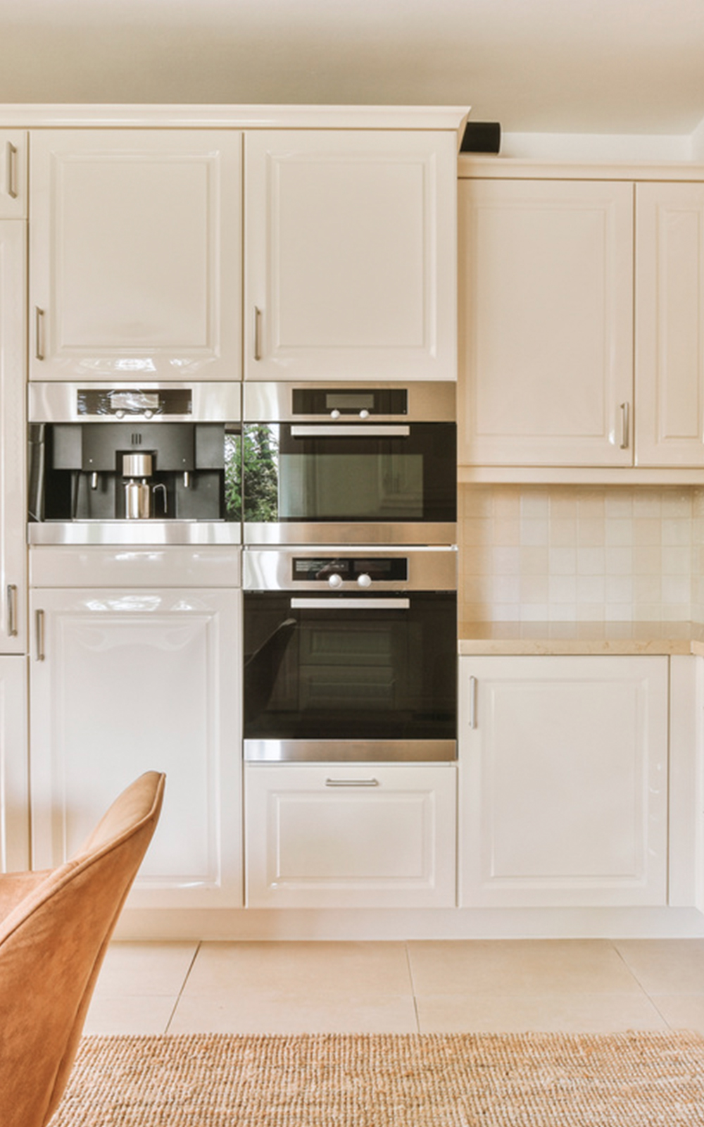 Modern kitchen design with glossy white cabinets, built-in stainless steel appliances including an oven and coffee machine, beige tiled floor, and a brown upholstered chair in the foreground.