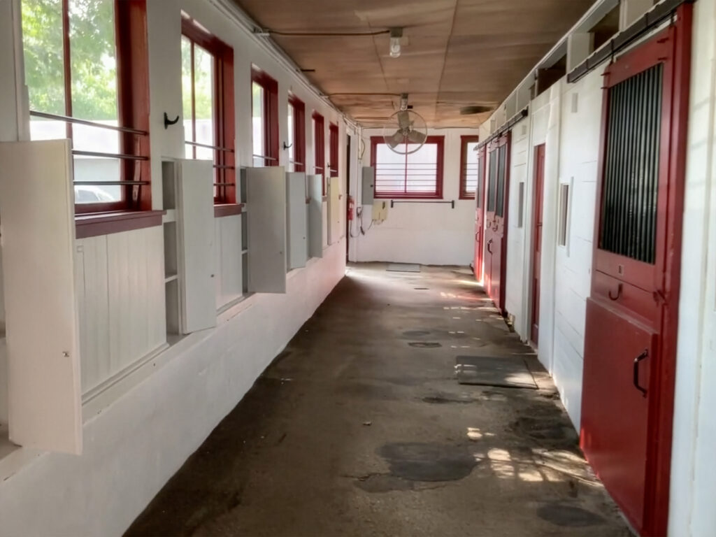 A clean hallway inside a stable with red and white stall doors on the right, open windows on the left, and sunlight streaming in from outside, resembling the tranquil light of a church painting.