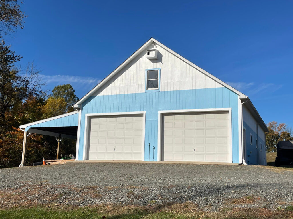 A light blue and white building with two large garage doors, a single window above them, and a carport on the left side sits on a gravel driveway under a clear blue sky. Trees are visible in the background, reminiscent of a peaceful church painting.