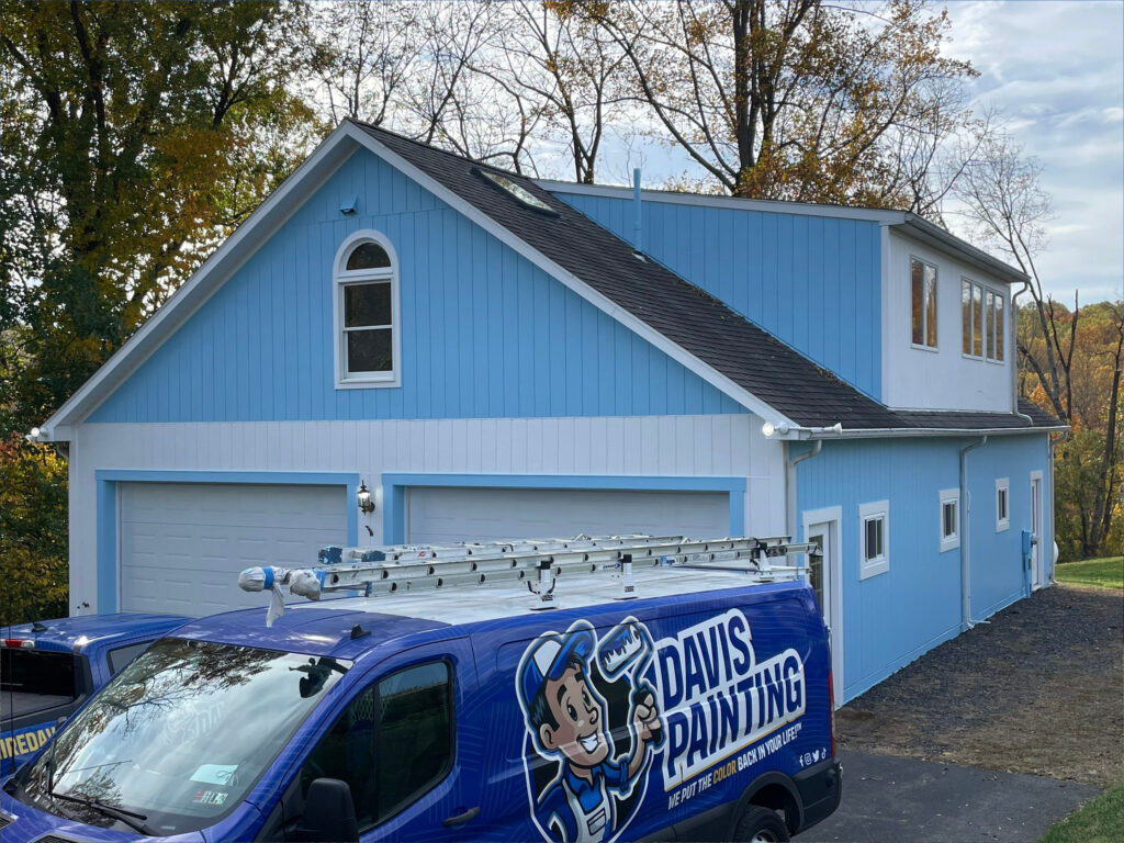 A light blue house with white trim and a detached garage is pictured. A blue van labeled "Davis Painting"—known for expert home and church painting—is parked in the driveway. Trees with autumn foliage are in the background.