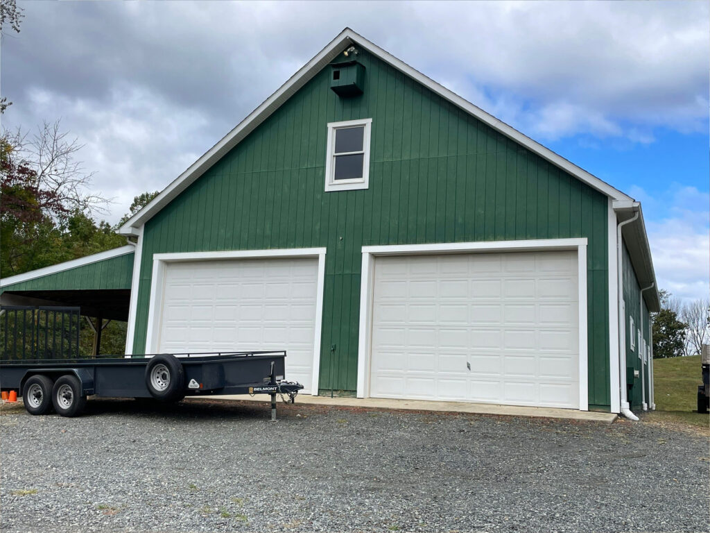 A green barn-style building with two large white garage doors, a small upper window, and a black utility trailer parked out front on a gravel driveway—its look reminiscent of church painting. Trees and a cloudy sky form the backdrop.