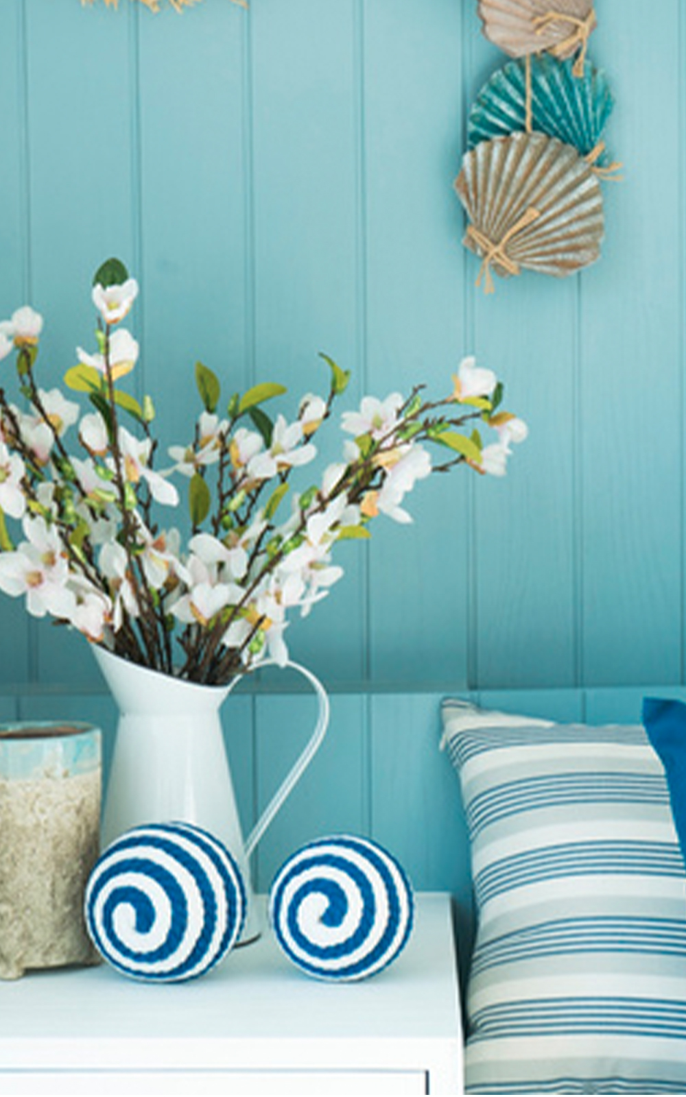 A white pitcher filled with blooming branches sits on a white table beside two blue and white spiral-patterned balls, a candle, and a striped pillow, against a light blue paneled wall with shell decorations that enhance the coastal design.