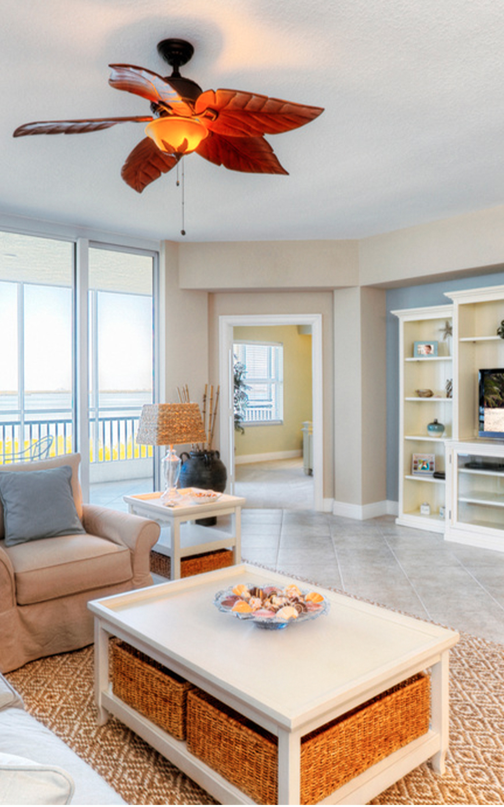Bright living room with large windows, a ceiling fan with leaf-shaped blades, white furniture, and built-in shelves. The elegant design features a coffee table with seashells, tiled floor, beige walls, and a patio with water view outside.
