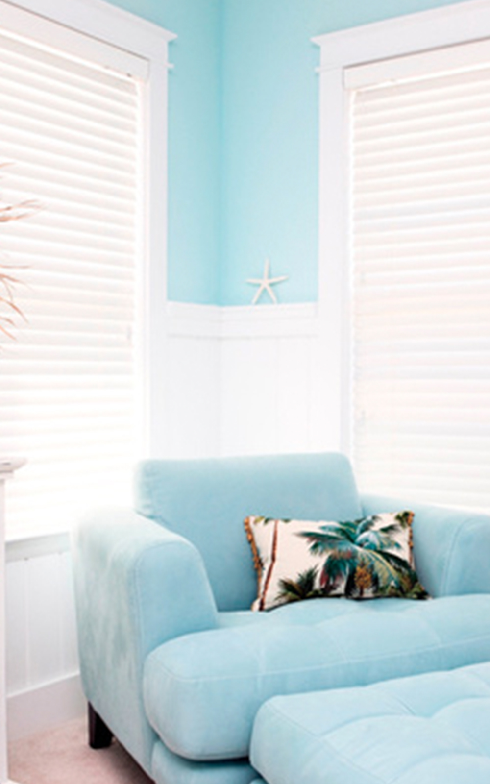 A light blue armchair with a tropical palm tree pillow sits in a bright room with sky-blue walls, showcasing coastal design. White blinds cover two windows, while a starfish decorates the windowsill in the corner.