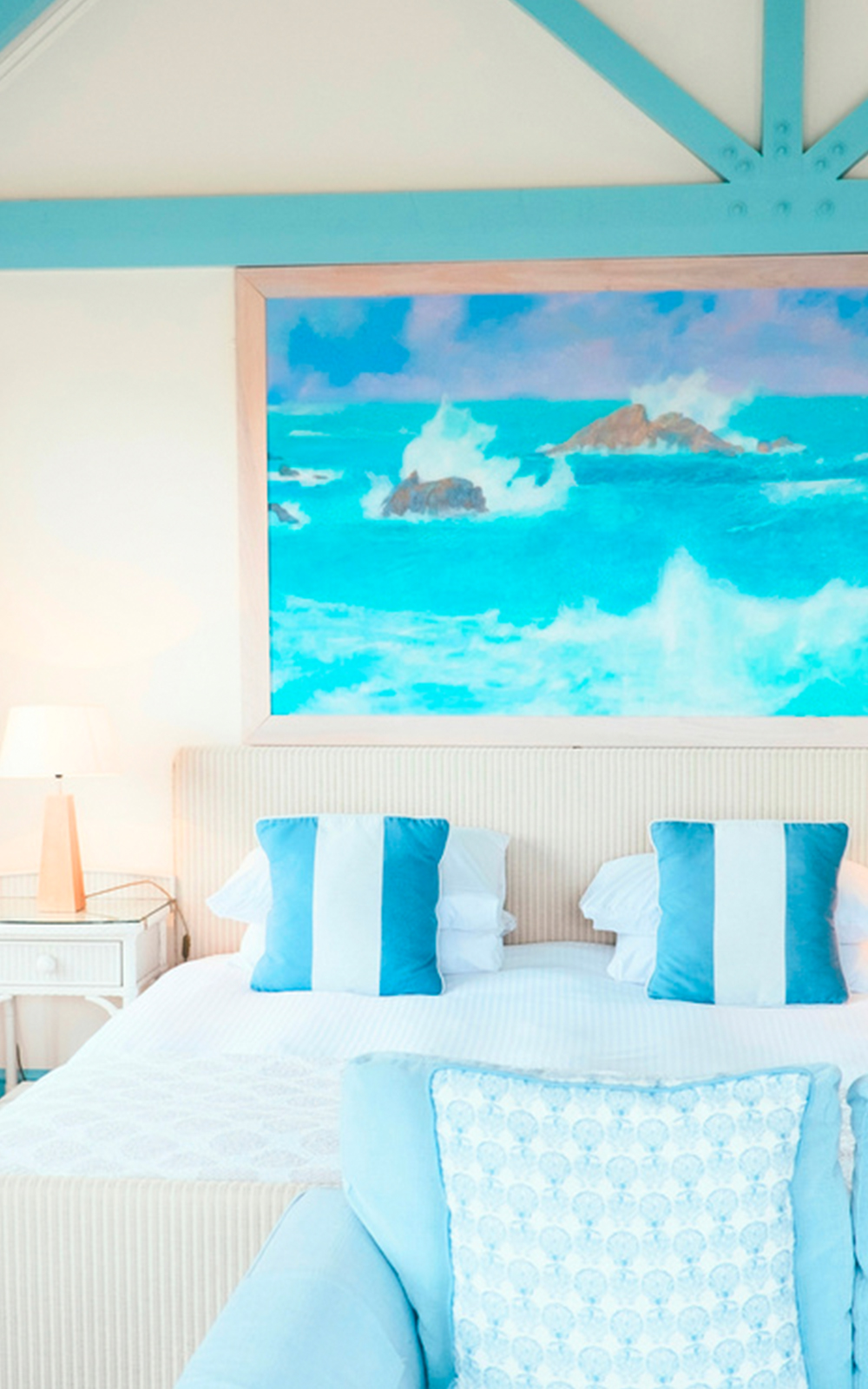 A bright, airy bedroom with a double bed featuring blue and white striped pillows, a light blue armchair, a bedside table with a lamp, and a large ocean-themed painting above the bed showcase thoughtful design elements.