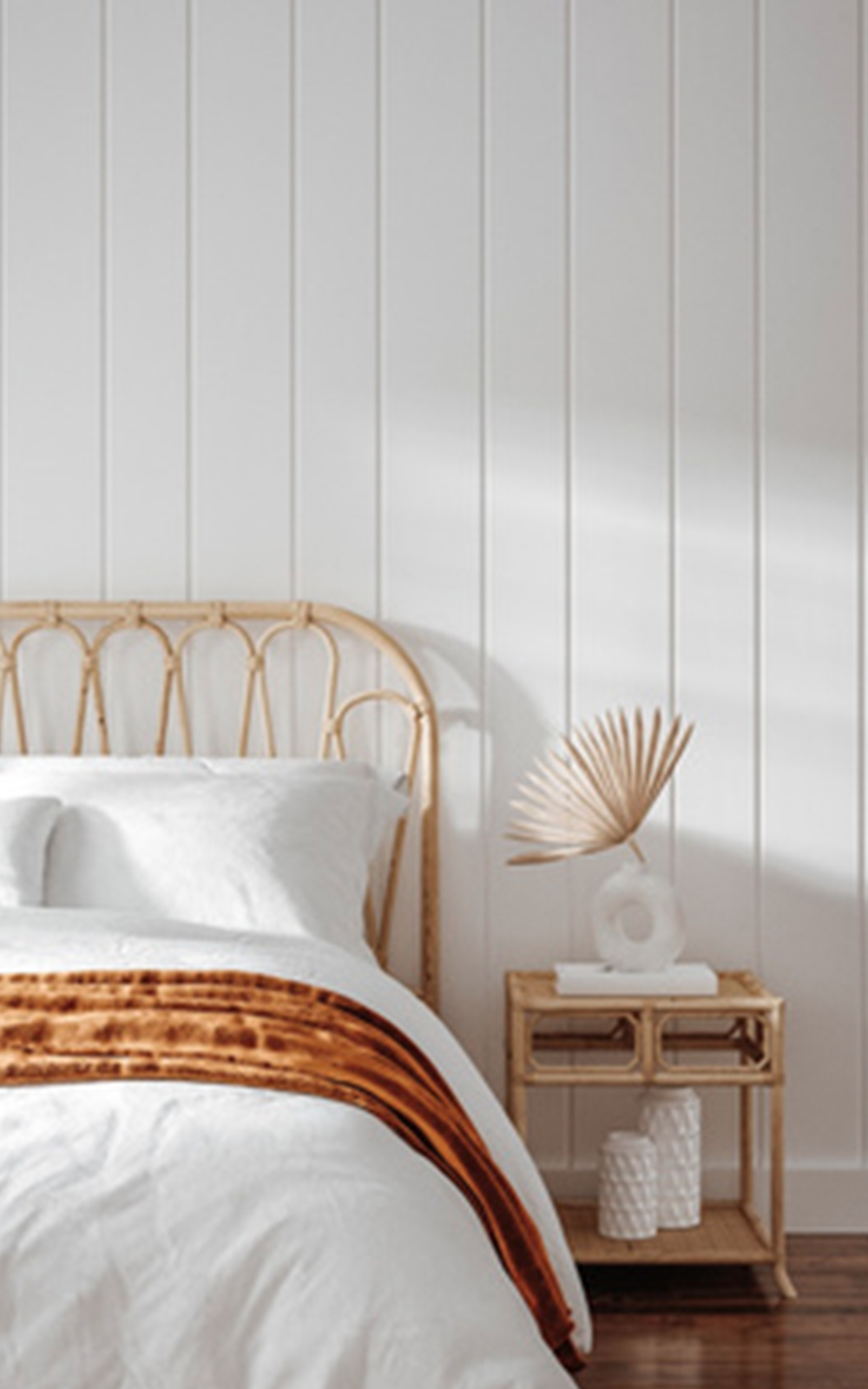 A cozy bedroom with a rattan headboard, white bedding, a brown throw blanket, and a wooden nightstand holding a decorative sculpture and dried palm leaf showcases thoughtful design against a white paneled wall.