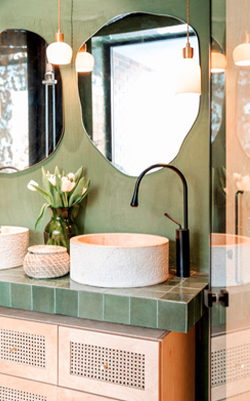 A stylish bathroom design featuring a green tiled countertop, two round mirrors, a white stone sink, a black faucet, a vase of white tulips, pendant lights, and wooden cabinets with rattan panels.
