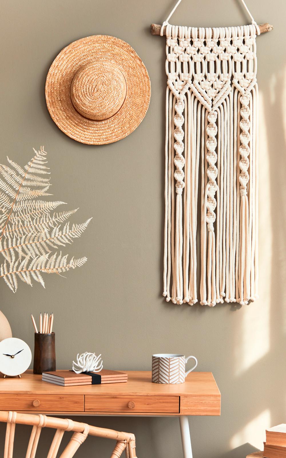 A cozy workspace with a wooden desk, clock, mugs, pencils, and books. Above the desk, a straw hat and macramé wall hanging add design flair to the sage green wall, where a leafy shadow is gracefully cast.