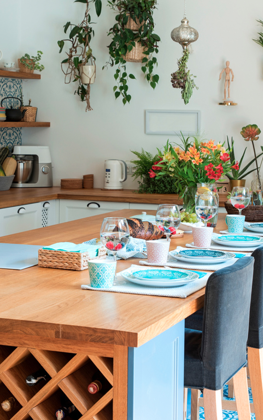 A bright, modern kitchen with thoughtful design features a wooden island set for a meal, blue patterned plates, cups, flowers, wine glasses, hanging plants, and open shelving showcasing kitchen appliances and decor.