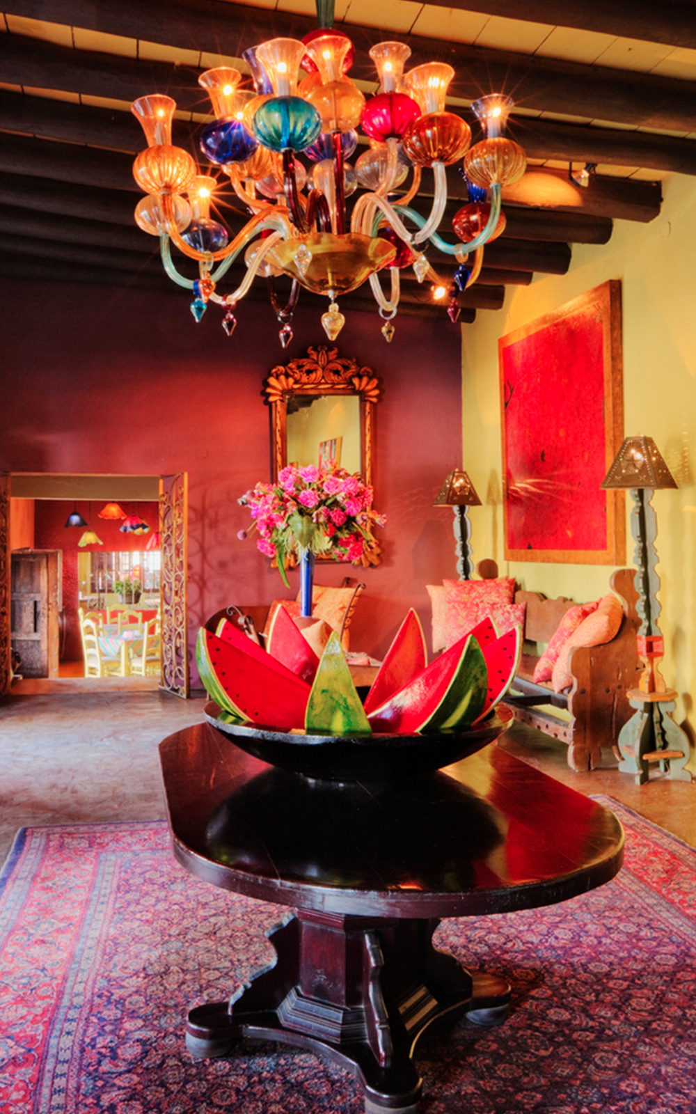 A vibrant room with colorful walls, a multicolored glass chandelier, design-forward eclectic furniture, a dark wood table holding a watermelon centerpiece, a bouquet of flowers, and a patterned rug.