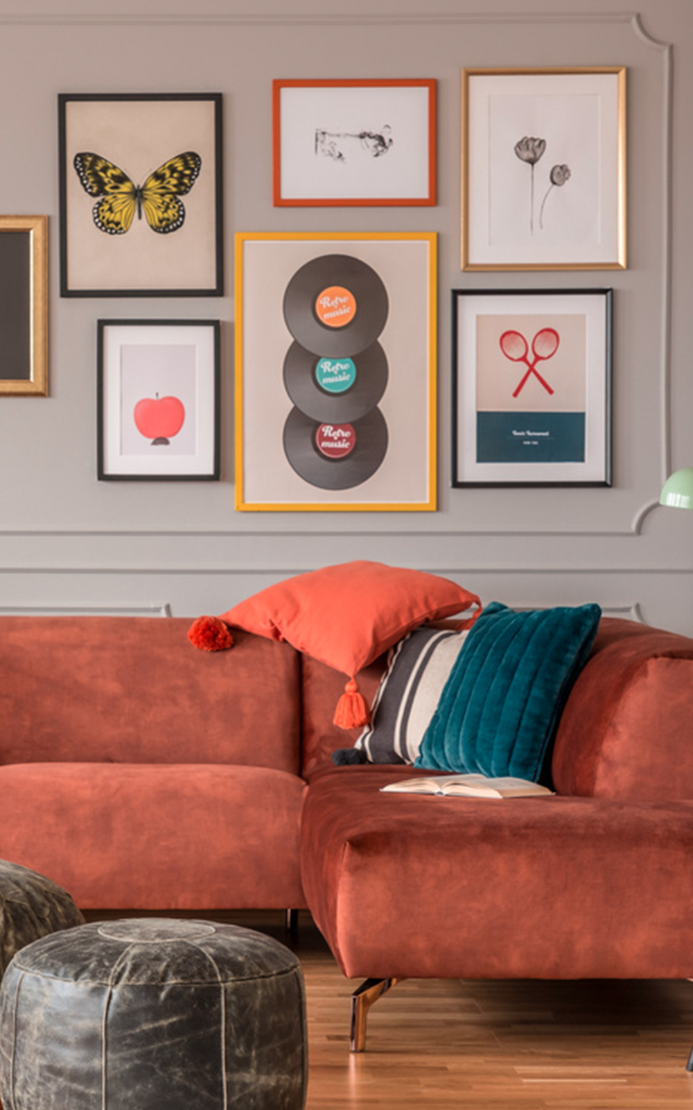 A cozy living room with a rust-colored sectional sofa, teal and orange pillows, and a gallery wall featuring framed artwork in a thoughtful design, including a butterfly, records, and simple illustrations. Two leather ottomans sit on a wooden floor.