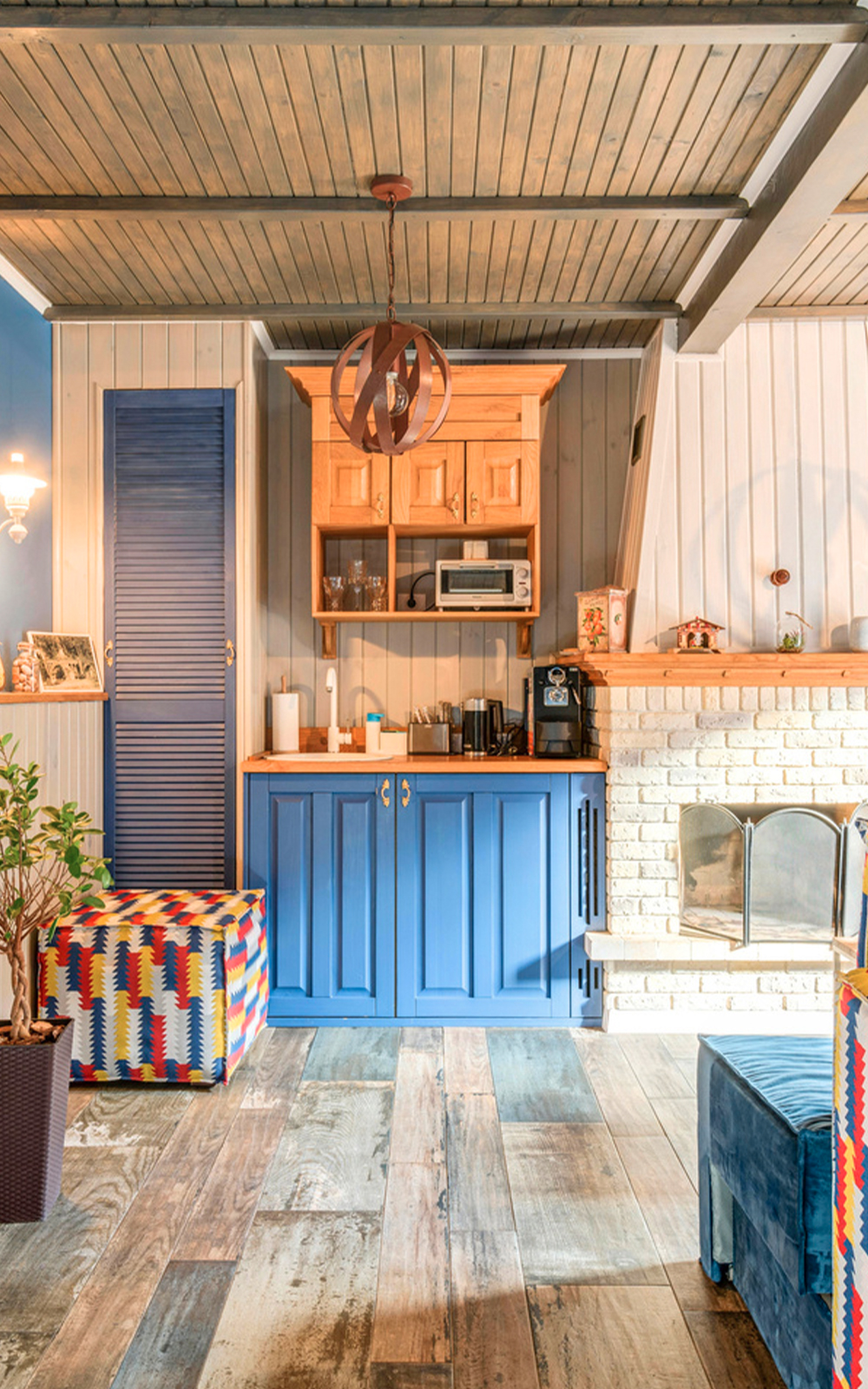 A cozy kitchen with blue cabinets, a wooden floor, and a fireplace with a white brick surround showcases thoughtful design, complemented by colorful patterned ottomans in a well-lit, rustic room with wooden ceiling beams.