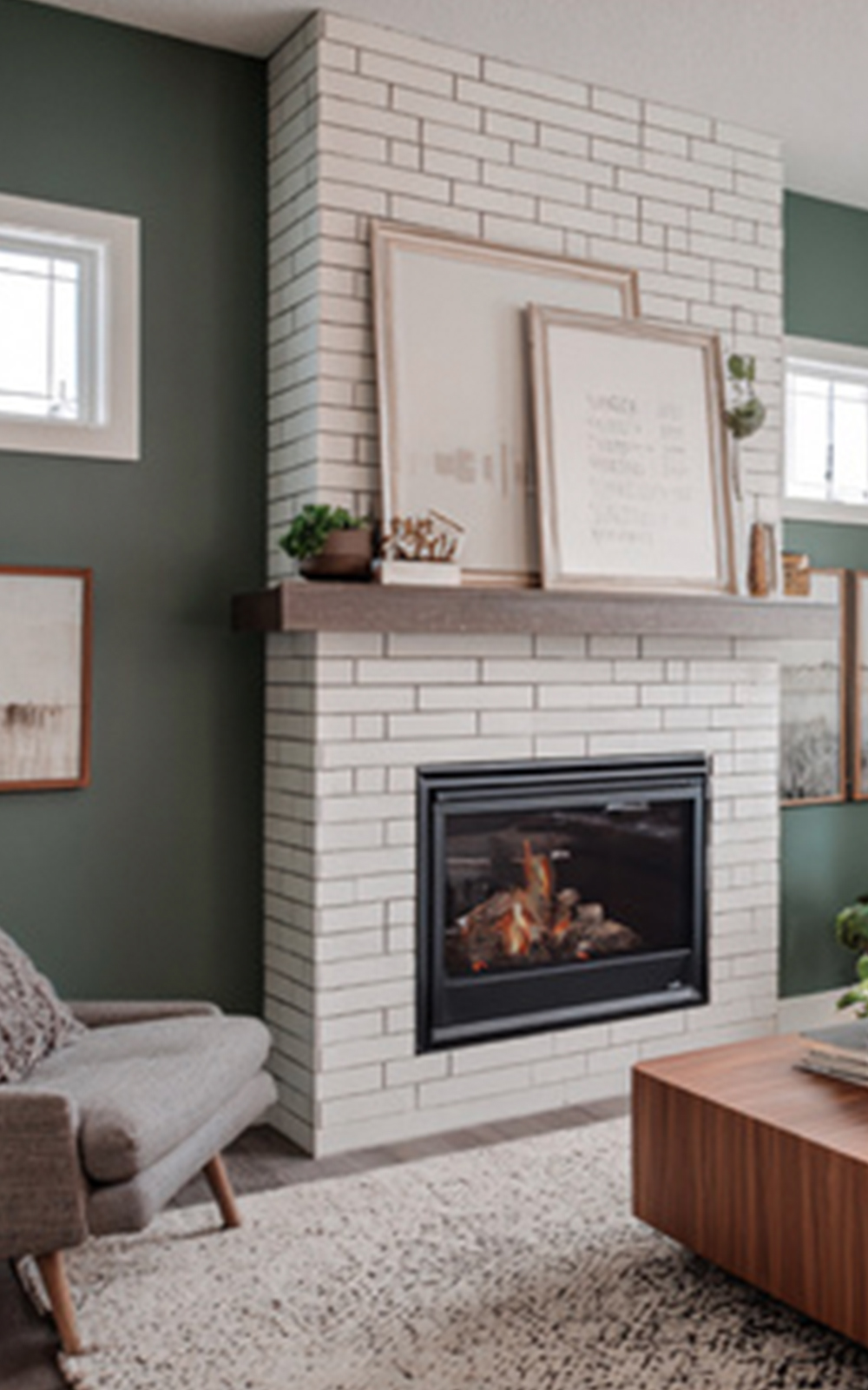 A modern living room with a white brick fireplace, gray armchair, wooden coffee table, and stylish design touches like framed art and plants on the mantel. The green walls add to the cozy, inviting atmosphere.