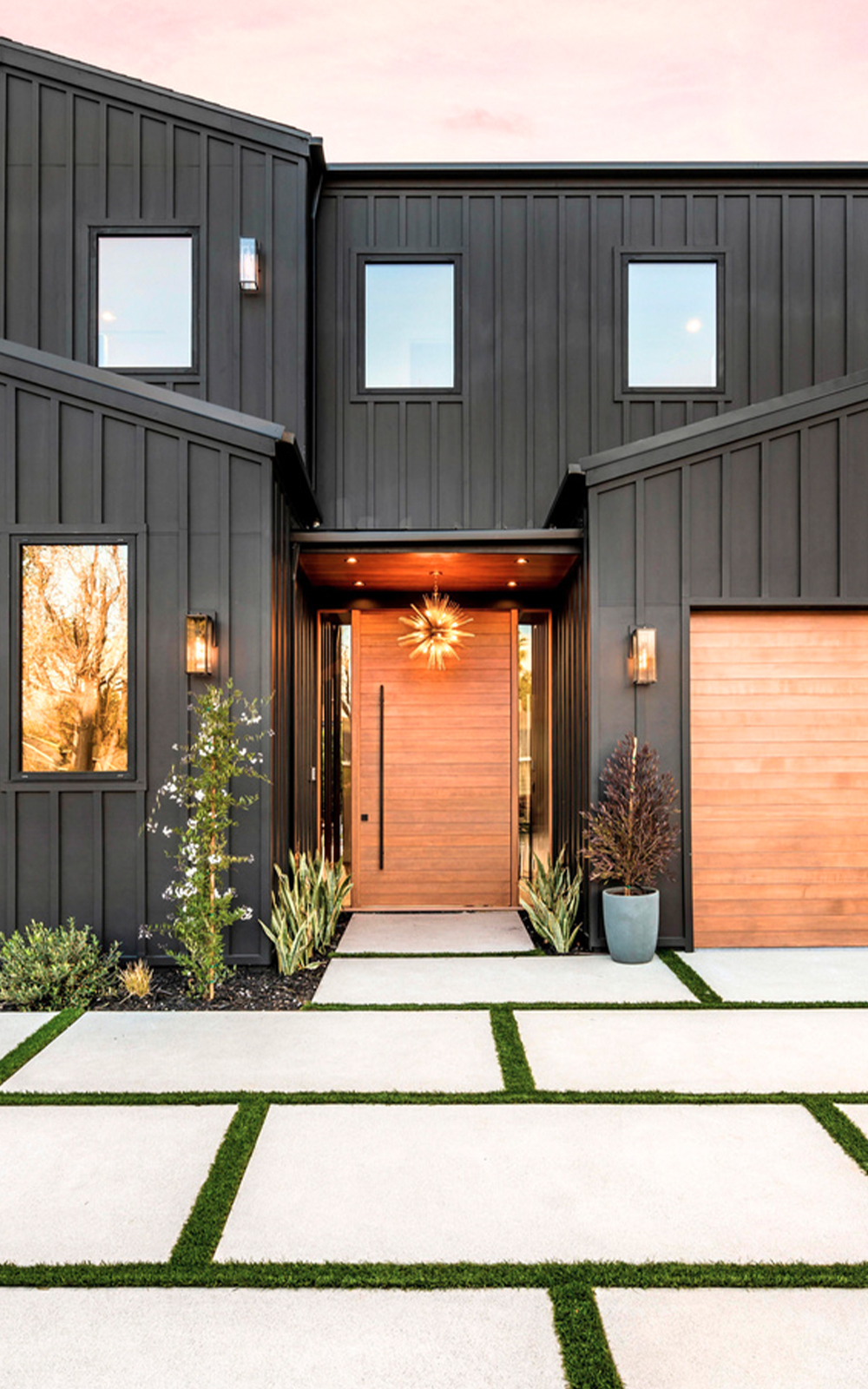 Modern two-story house with black siding and large windows showcases a sleek design. A wooden front door and matching garage door complement the geometric concrete pavers and green grass strips in the minimal front yard landscaping.