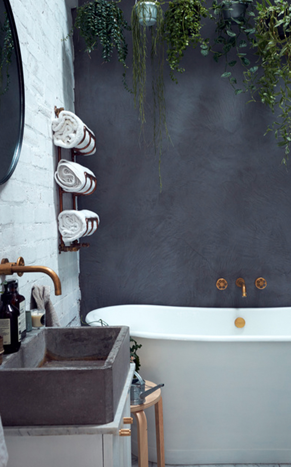 A stylish bathroom with a gray wall, white bathtub, brass fixtures, and a concrete sink showcases thoughtful design. Towels rolled on a metal rack, green plants hanging above, and a round mirror on a white brick wall complete the look.