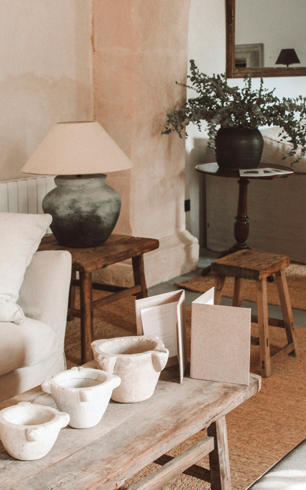 A cozy living room with a neutral palette features a beige sofa, rustic wooden tables, ceramic pots, and thoughtful design touches like a dark-base lamp, potted plant, and wicker rugs, creating a warm and inviting atmosphere.