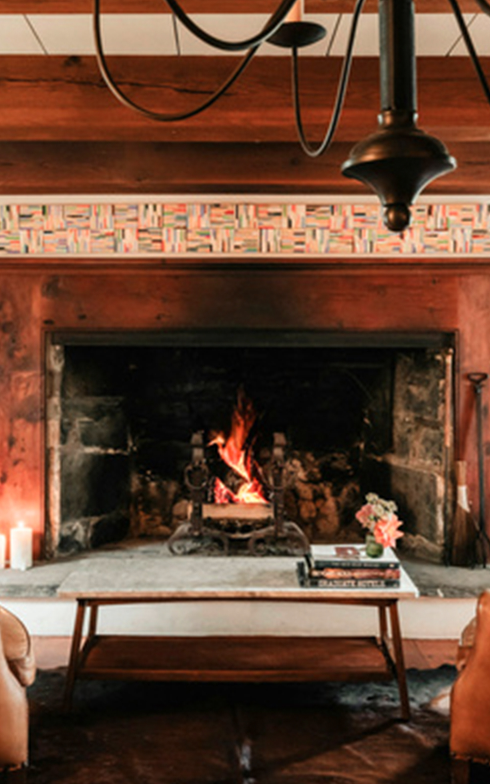 A cozy living room with a lit fireplace showcases warm design touches—a wooden mantle decorated with colorful books, a coffee table with books and flowers, leather chairs, and a candle on the hearth.