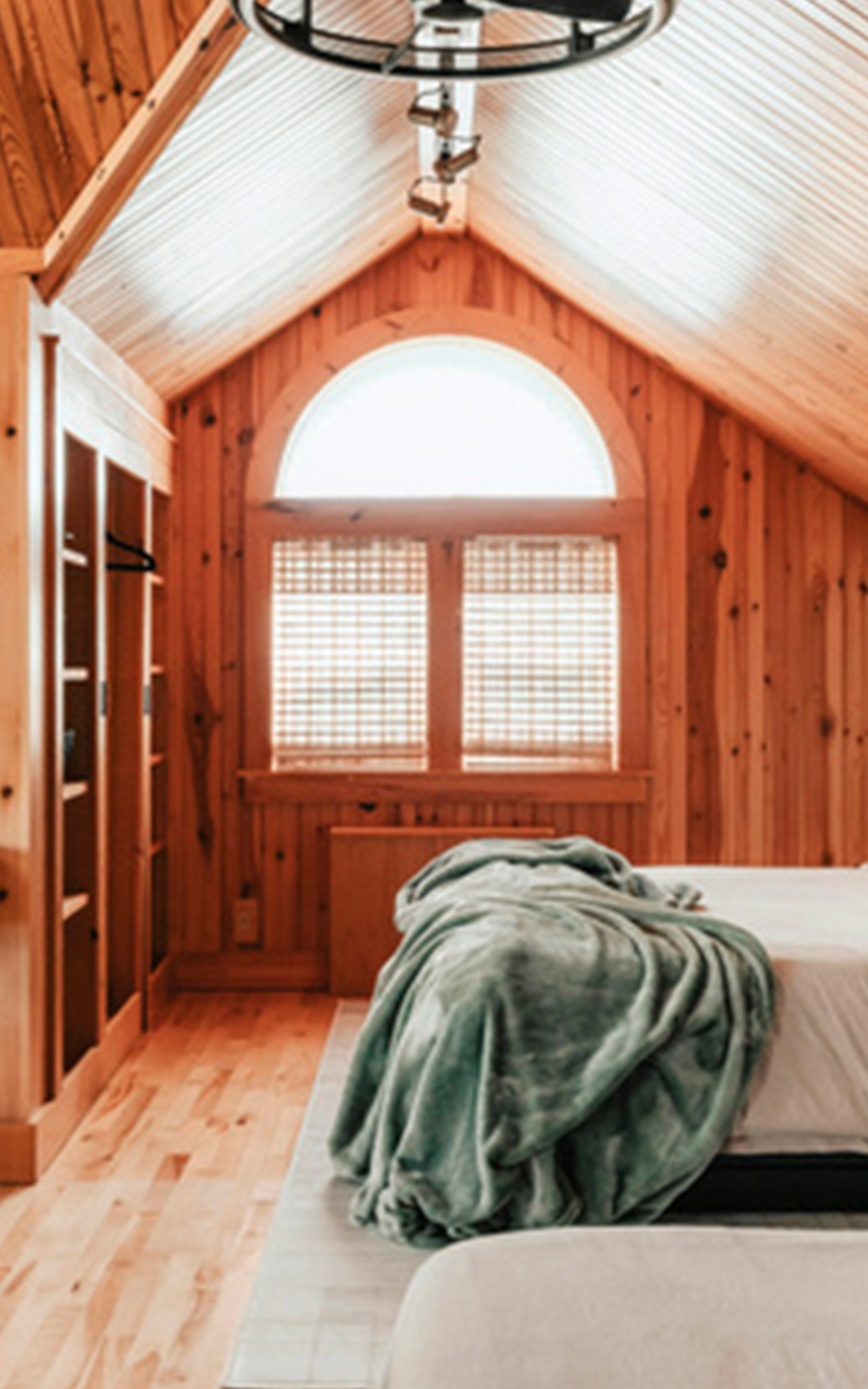 A cozy wooden attic bedroom with a sloped ceiling, arched window, and thoughtful design features. Two beds with white linens and a green blanket rest beneath sunlight filtering through blinds, highlighting the natural wood interior.